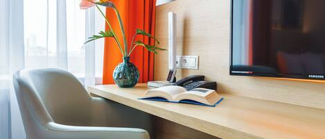 A hotel room with a wooden desk, a modern TV, a vase with flowers, and a city view.