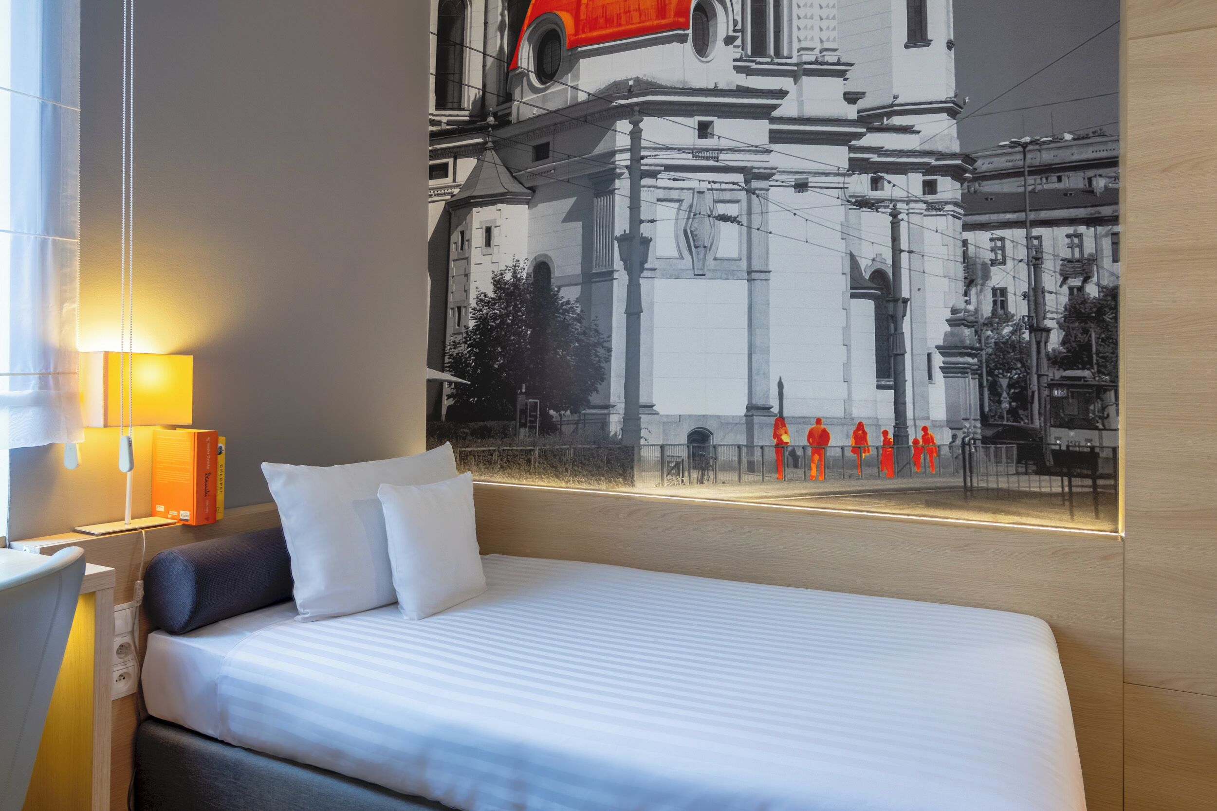 Une chambre d’hôtel avec un grand lit, une table de chevet, une lampe et une fresque murale représentant un bâtiment.