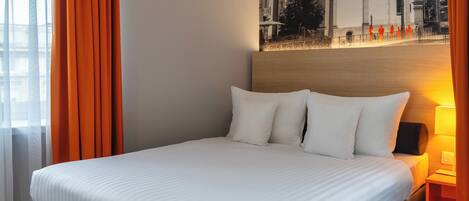A neatly made bed with white linens and pillows, a wooden headboard, and a wall-mounted lamp.