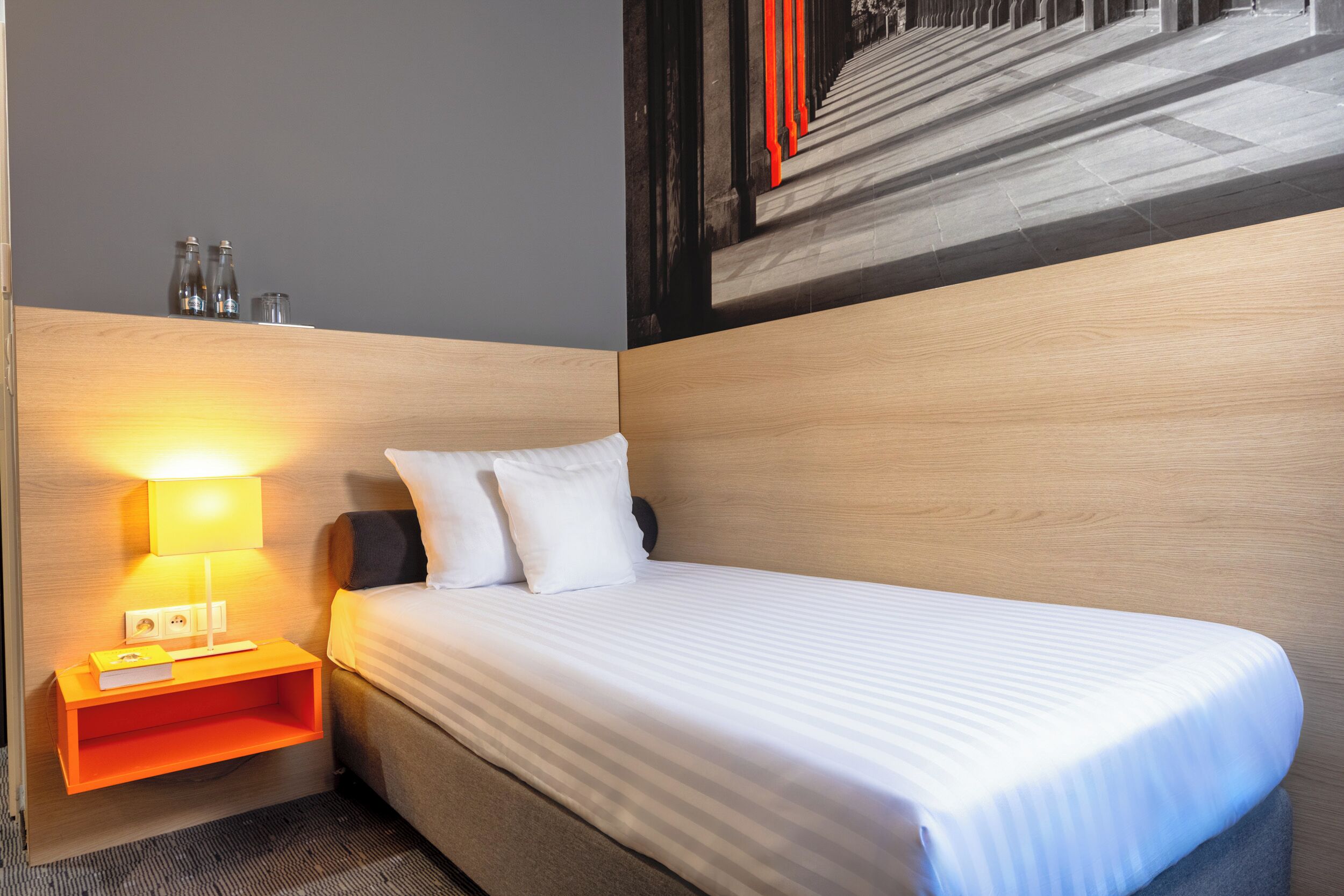 Une chambre d’hôtel avec un lit, une table de chevet avec une lampe et une étagère fixée au mur avec des verres.