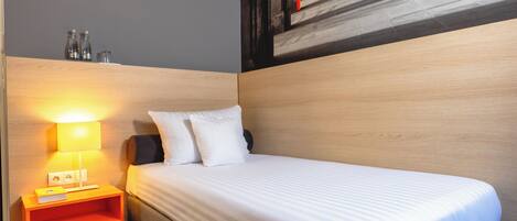 Habitación de hotel con cama, mesita de noche con lámpara y estante en la pared con vasos.