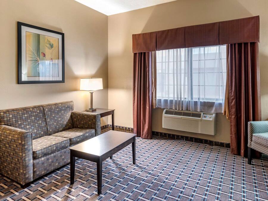 A hotel room with a patterned floor, a sofa, a small table, a lamp, a window with curtains, and an air conditioning unit.