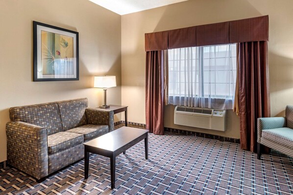 A hotel room with a patterned floor, a sofa, a small table, a lamp, a window with curtains, and an air conditioning unit.
