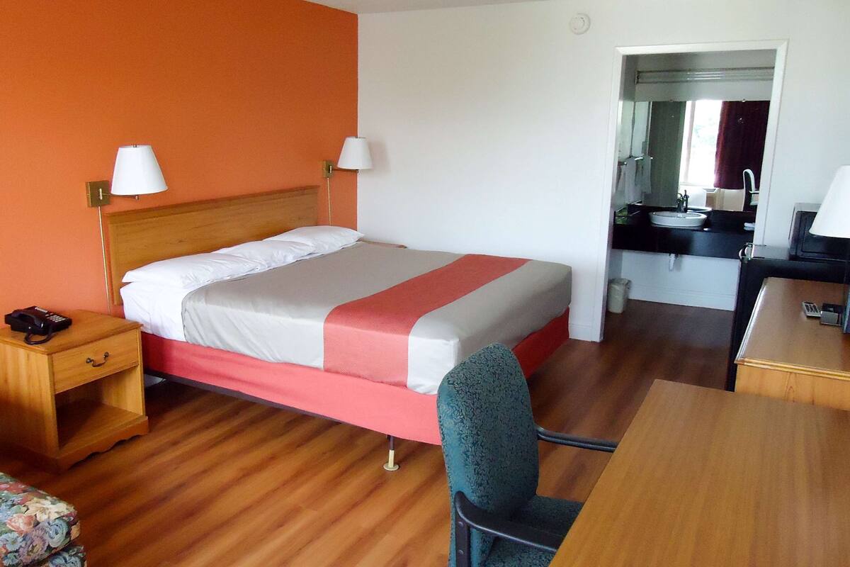 A hotel room with a bed, a desk, a chair, a nightstand, and a bathroom visible through an open door.