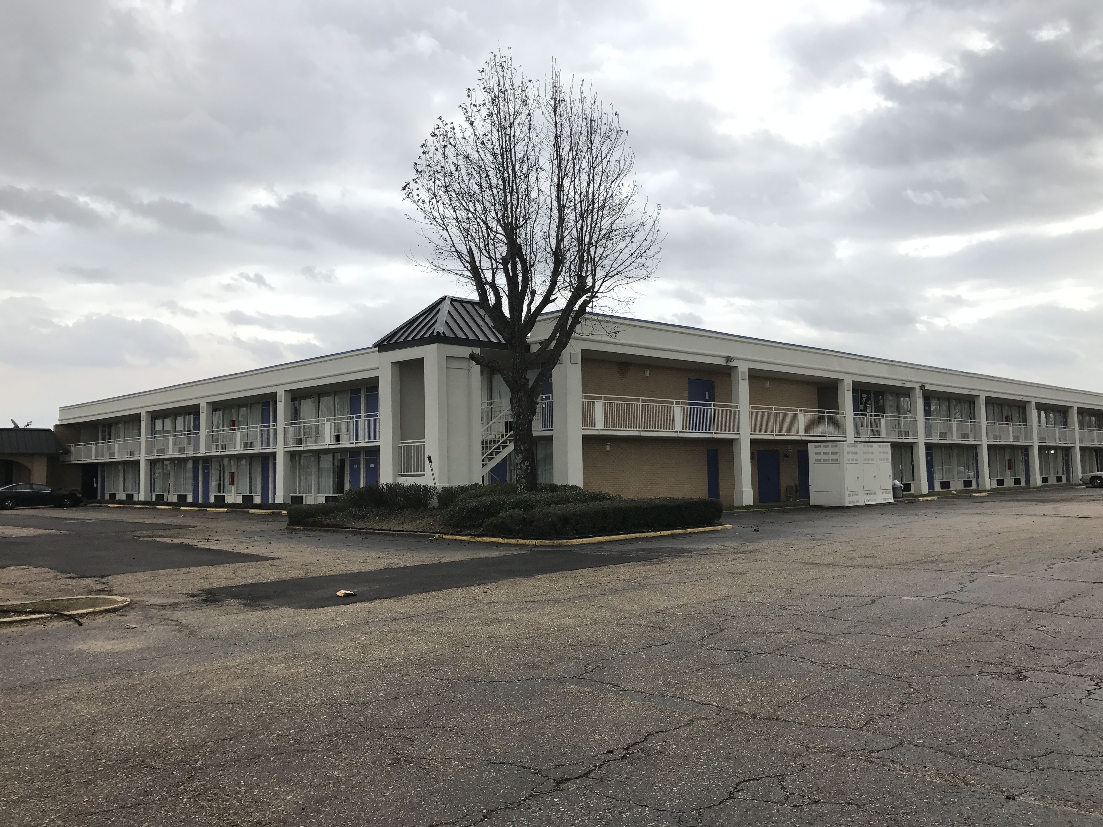 A multi-story motel with a central staircase, a parking lot, and a large tree in front.