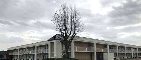 A multi-story motel with a central staircase, a parking lot, and a large tree in front.