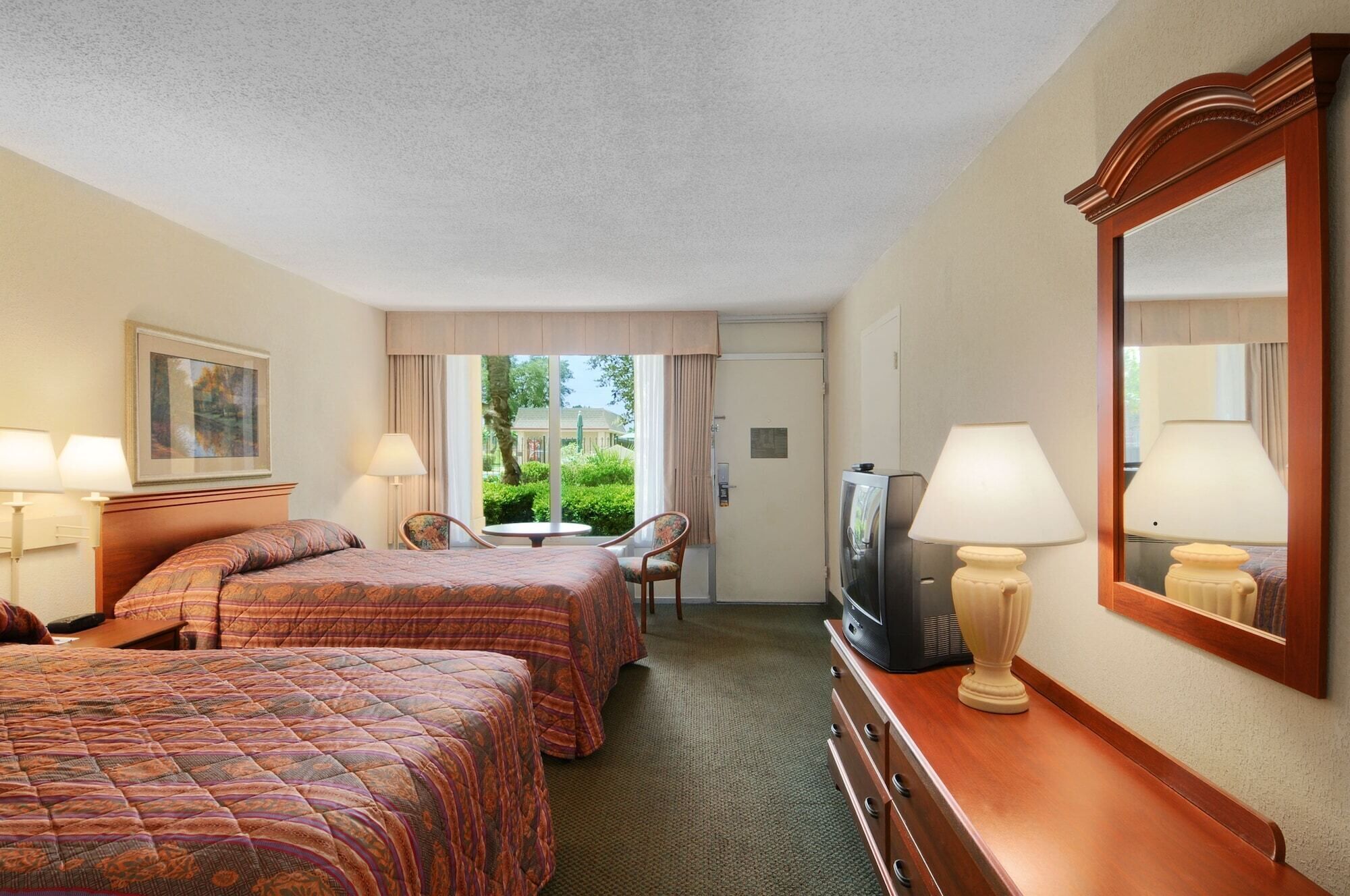 Ein Hotelzimmer mit zwei Betten, einem Schreibtisch mit Fernseher, einem Spiegel und einem Fenster mit Vorhängen.