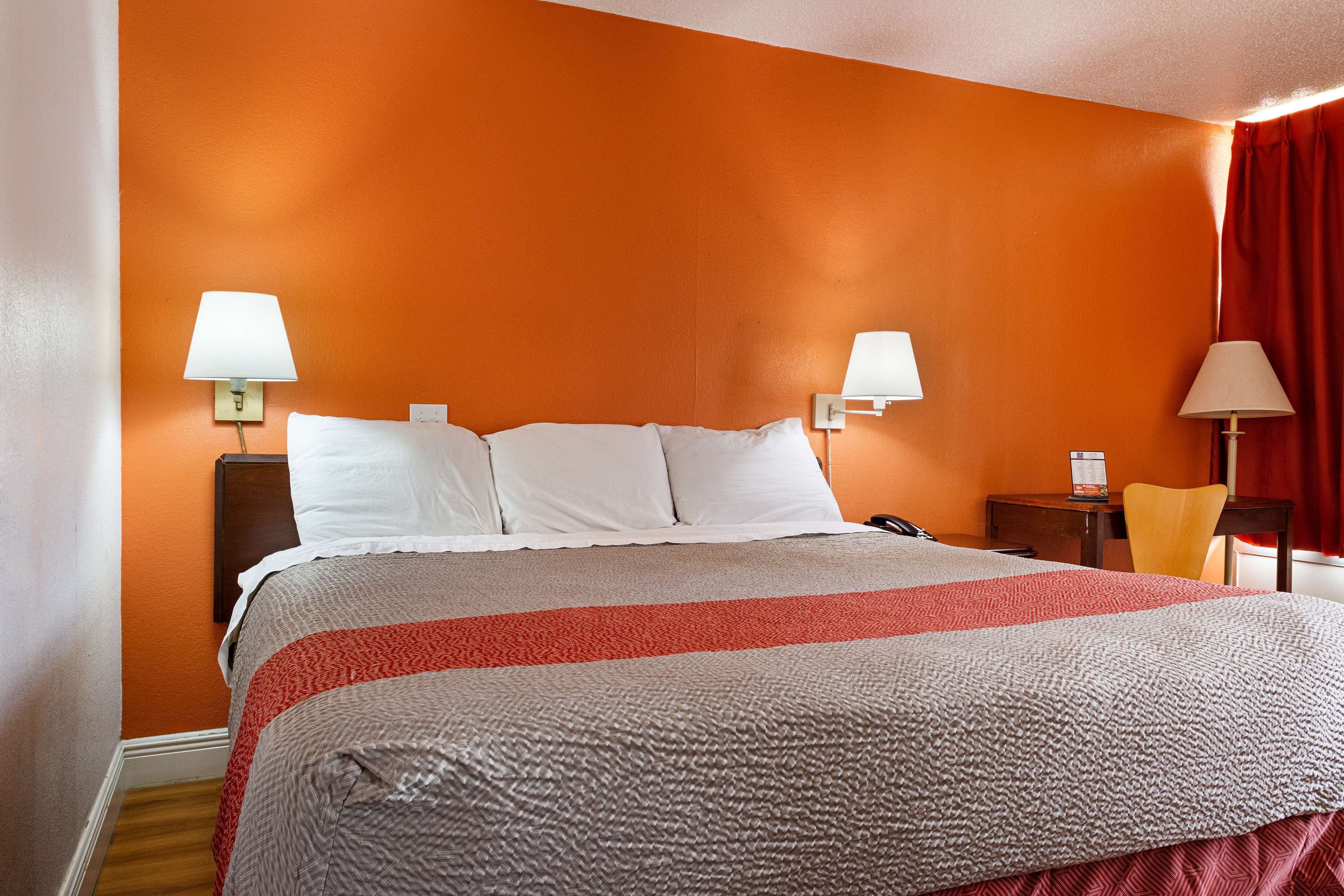 Une chambre d’hôtel avec un mur orange, un lit avec une literie blanche et rouge, deux lampes de chevet, une table de chevet en bois et un rideau rouge.