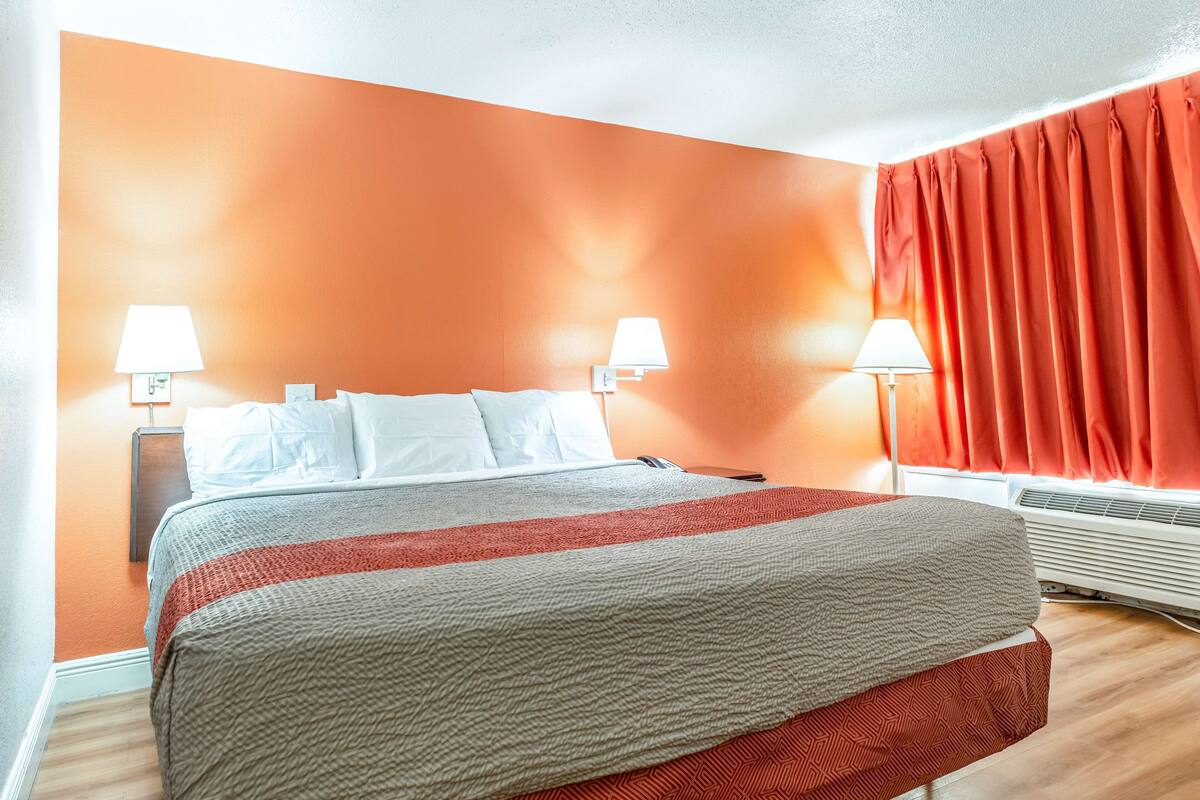 A hotel room with a bed, two bedside lamps, a window with curtains, and an air conditioning unit.