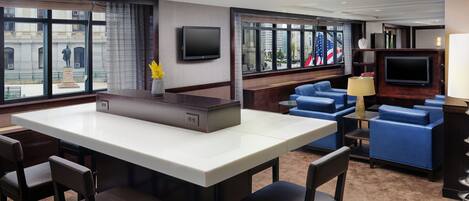 Un hall de hotel con una zona de bar central, asientos con cojines azules, un televisor de pantalla plana en la pared y grandes ventanales que ofrecen vistas de la ciudad.