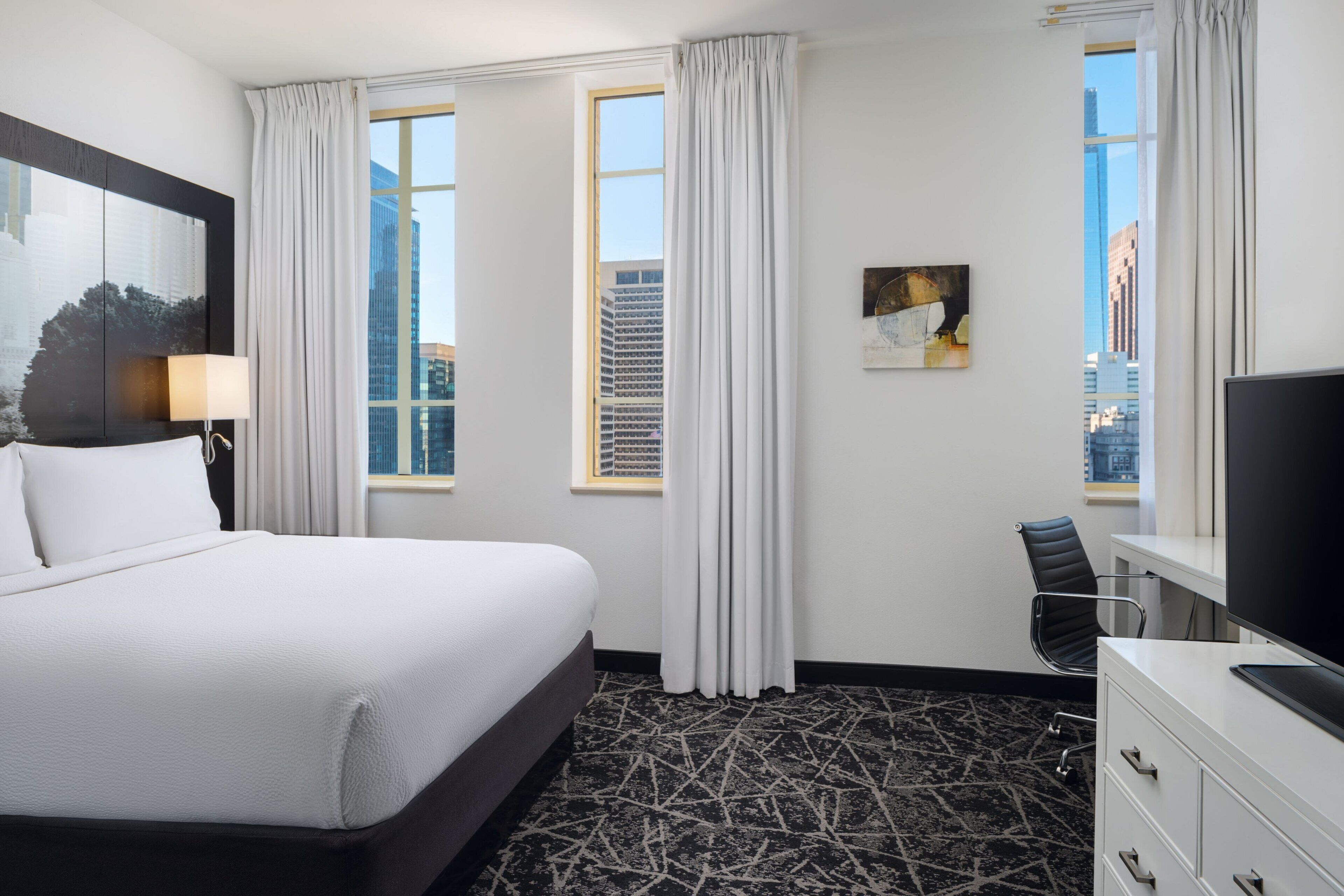 A hotel room with a large bed, a desk with a TV, a chair, and a painting on the wall.