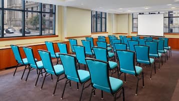 A conference room with rows of blue chairs, a white projection screen, and large windows with a city view.
