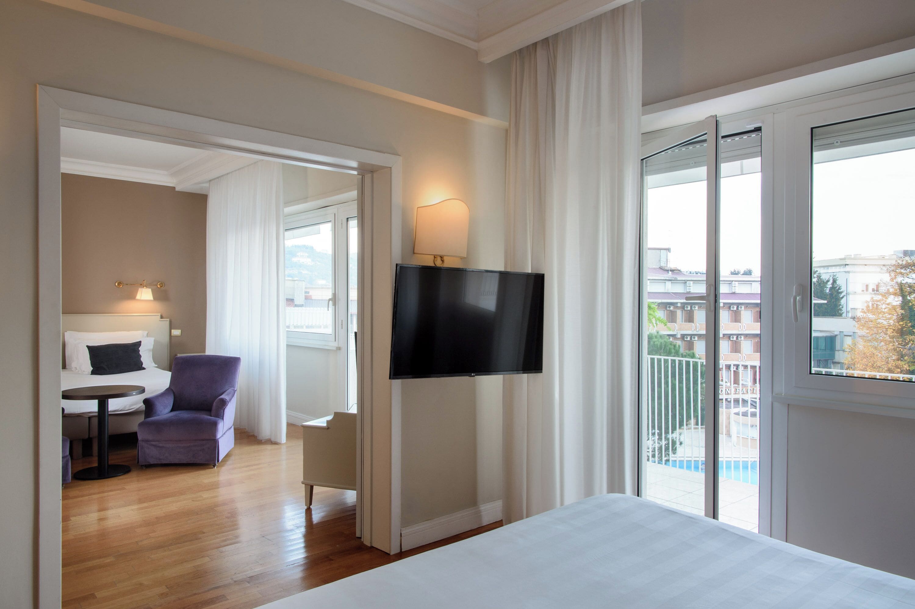 A hotel room with a bed, a TV mounted on the wall, a chair, a small table, and a balcony with a view of the city.