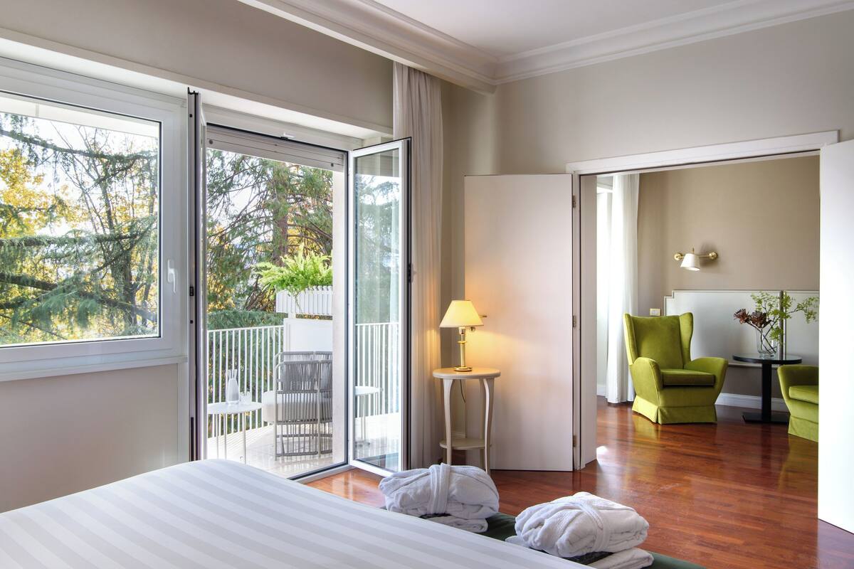 Habitación de hotel con cama, balcón con mesa y sillas, un sillón verde y una lámpara.