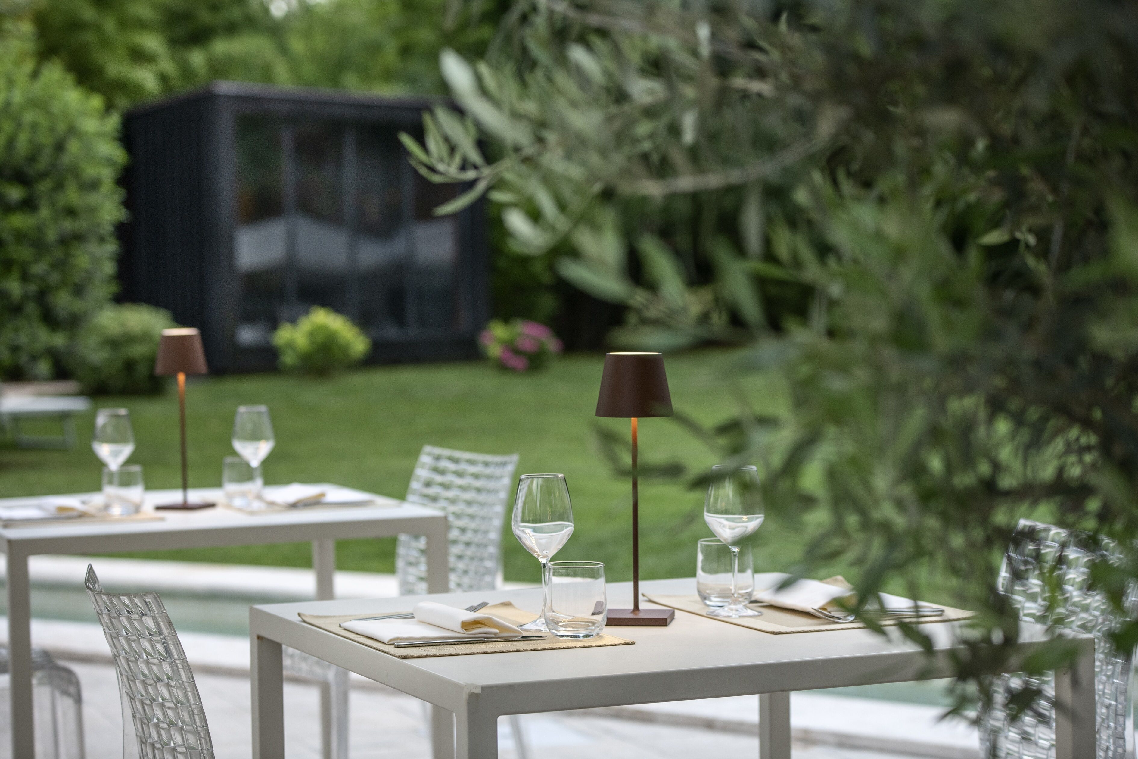 An outdoor dining area with tables set for two, each with a wine glass, a water glass, and cutlery.