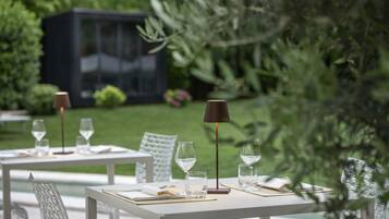 Un área de comedor al aire libre con mesas dispuestas para dos, cada una con una copa de vino, un vaso de agua y cubiertos.