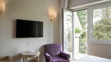 Ein Hotelzimmer mit Bett, einem an der Wand befestigten Flachbildfernseher, einem violetten Sessel, einem kleinen runden Tisch mit einem Glasbehälter und Gläsern sowie einem großen Fenster mit Blick auf Bäume und einen Balkon.