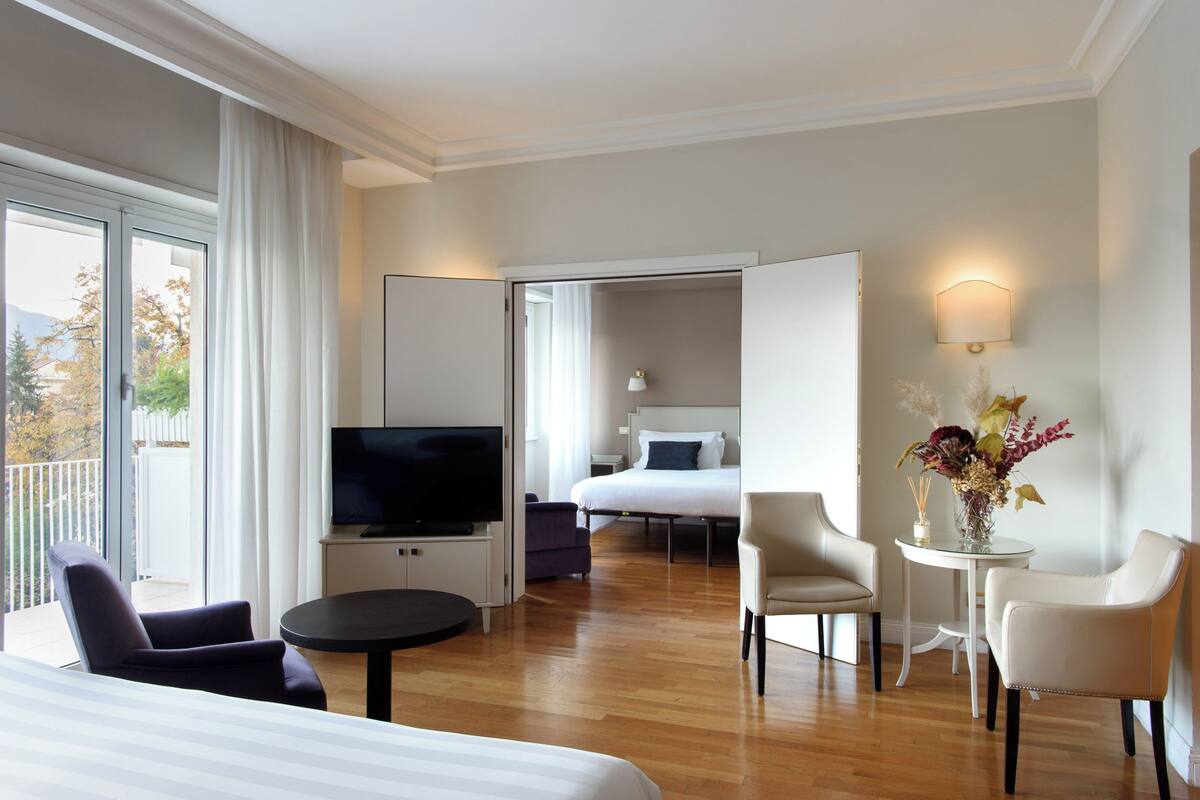 Habitación de hotel con cama, televisión, sofá, mesa de comedor y balcón con cortinas.