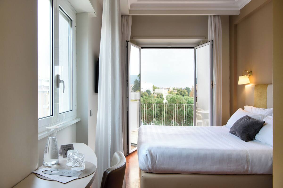 Habitación de hotel con cama, escritorio y silla, mesita con vaso, ventana y balcón con vistas a la vegetación.
