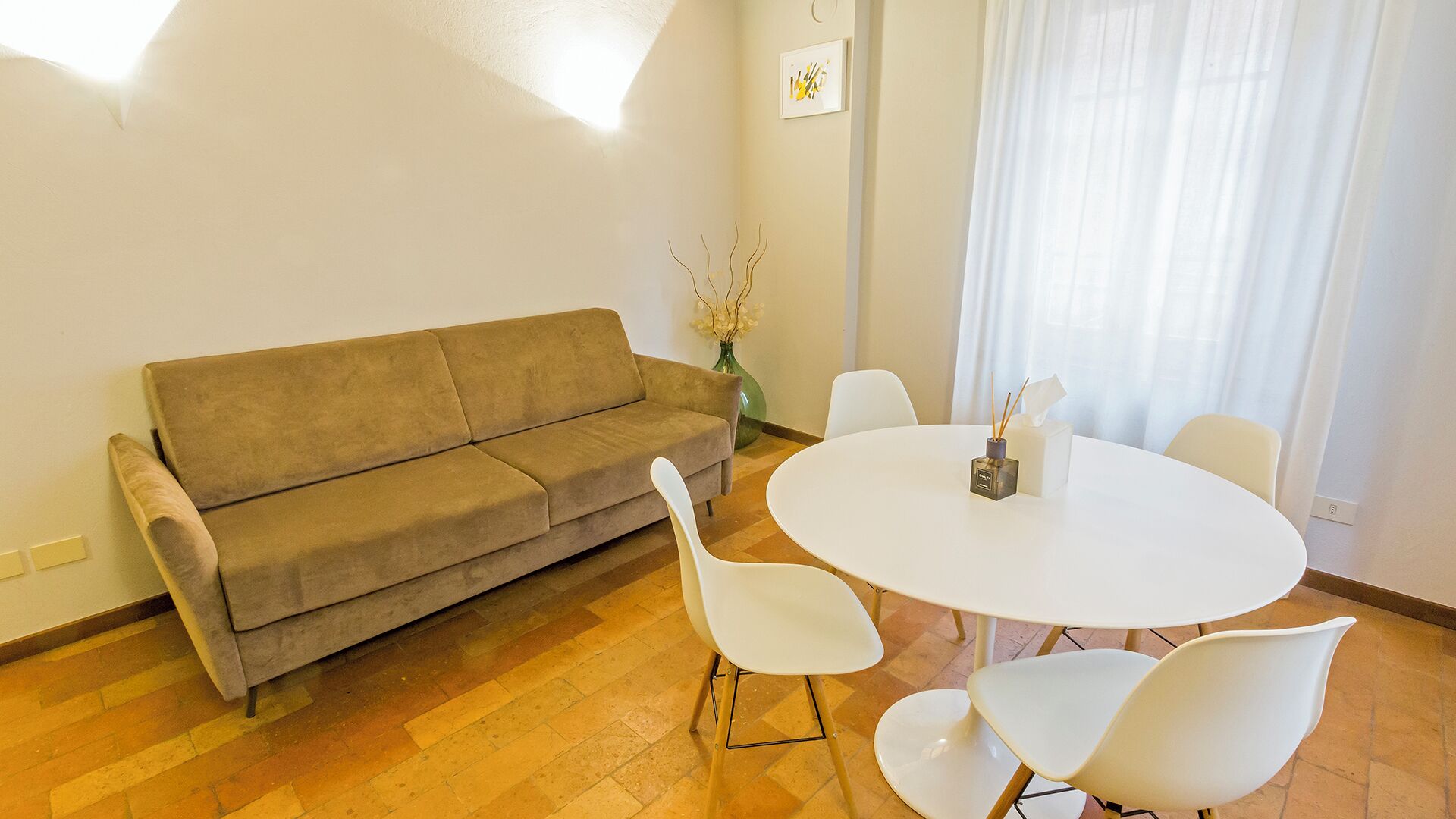 A living room with a beige sofa, a round white table, and four white chairs. There is a vase with flowers on the table and a picture on the wall.