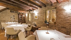 A restaurant with multiple round tables, each set with white tablecloths and chairs. There are wine glasses and a menu on the tables. The walls are made of exposed brick, and there is a wooden ceiling with beams.