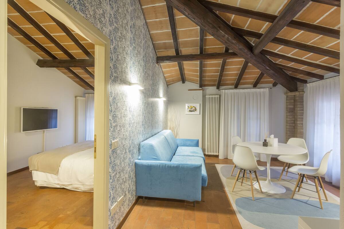 Un dormitorio con cama, sofá azul, mesa de comedor con sillas y techo con vigas a la vista.