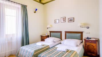 A hotel room with two beds, wooden flooring, a ceiling with decorative patterns, a window with sheer curtains, and framed pictures on the wall.