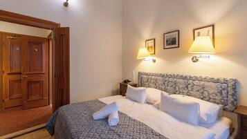 Una cama bien tendida con sábanas blancas y un cabecero estampado con motivos florales, dos lámparas de noche y una puerta de madera con cerradura.
