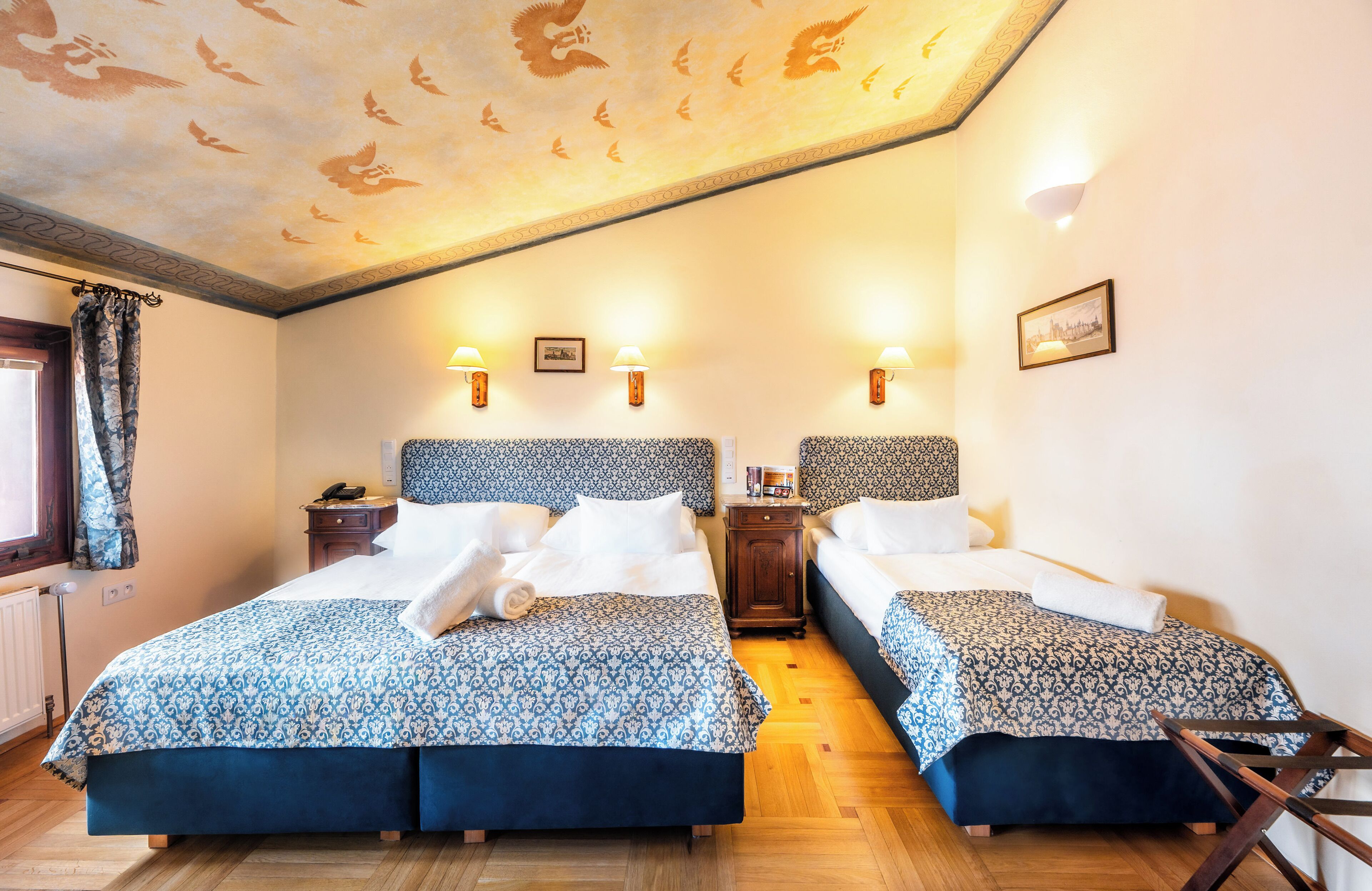 Habitación de hotel con dos camas, piso de madera, techo estampado y lámparas fijadas a la pared.