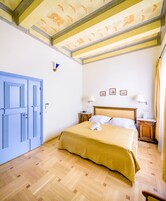 Quarto de hotel com cama, piso de madeira, porta azul e teto com padrão geométrico.