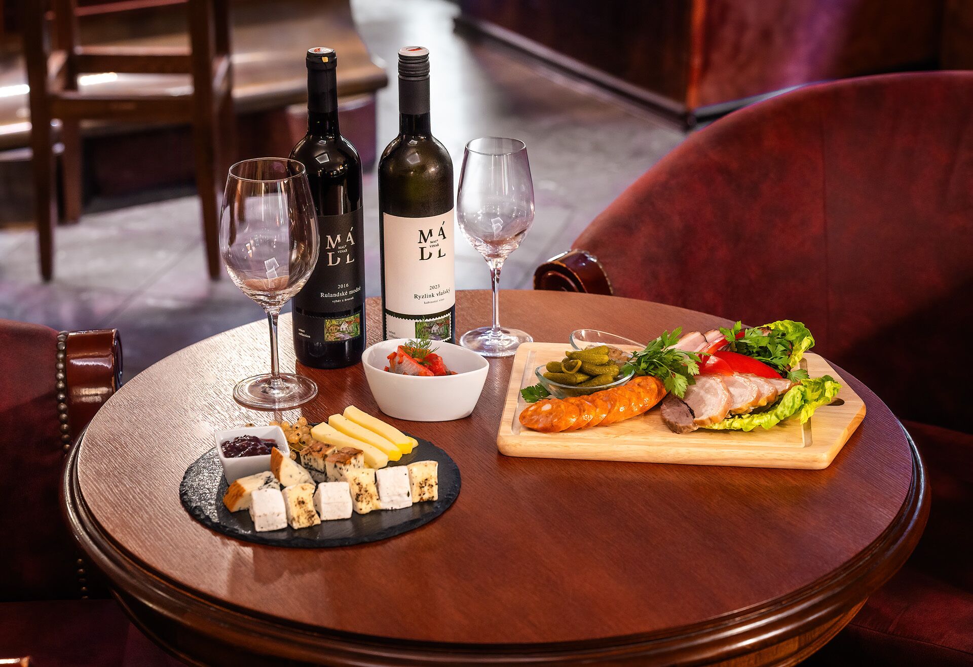 A round wooden table with a bottle of wine, two glasses, a bowl of cheese, and a wooden board with sliced meat and vegetables.