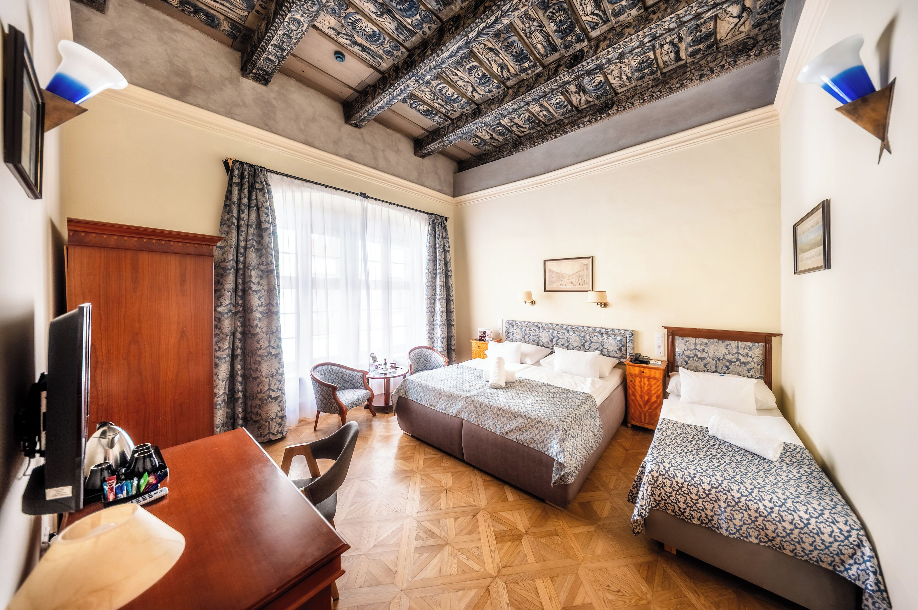 Une chambre d’hôtel avec un bureau en bois, deux lits, une télévision et un plafond orné de motifs complexes.