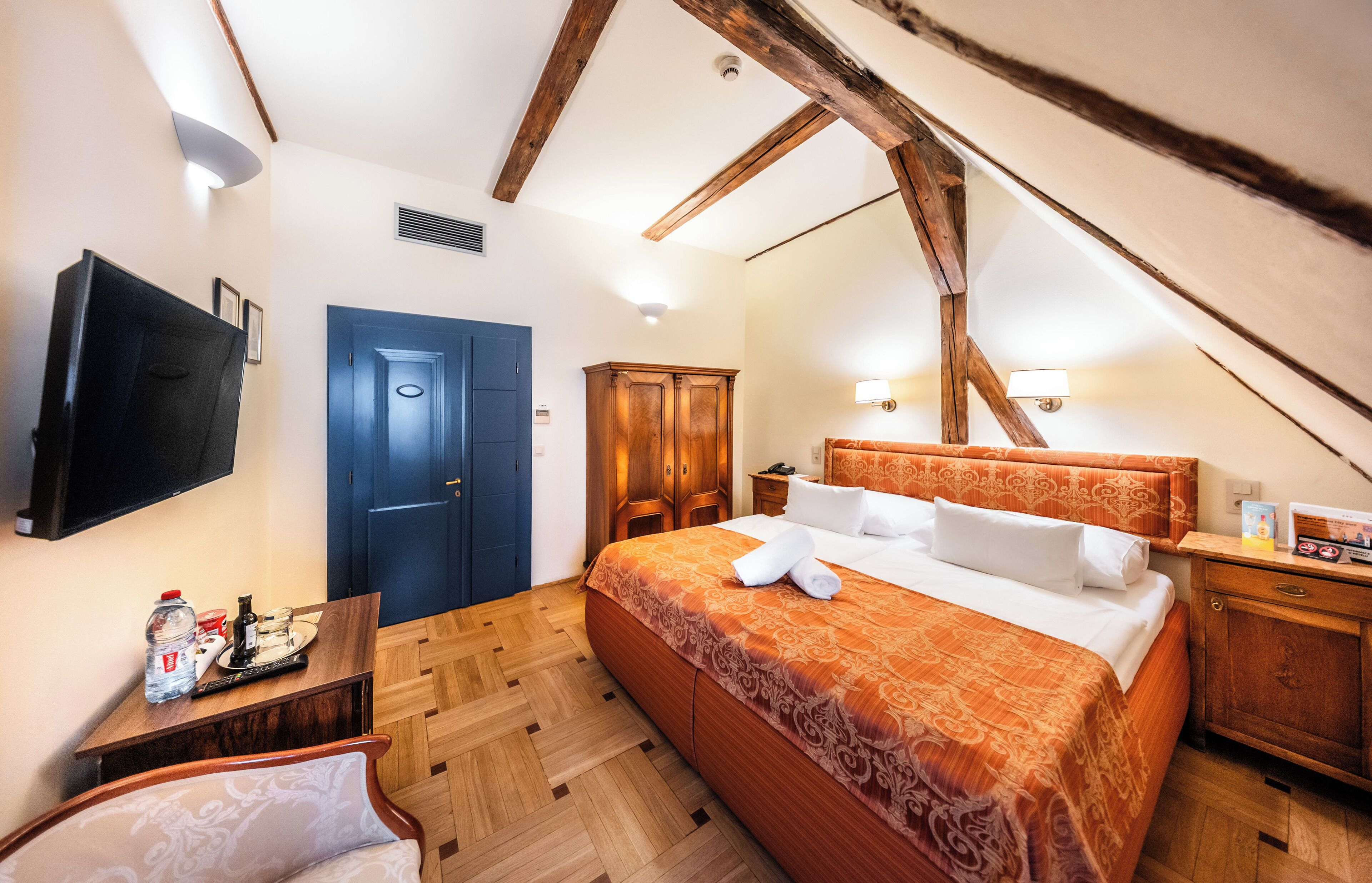 A bedroom with a wooden ceiling, a bed with an orange bedspread, a TV, a desk with a bottle, and a blue door.