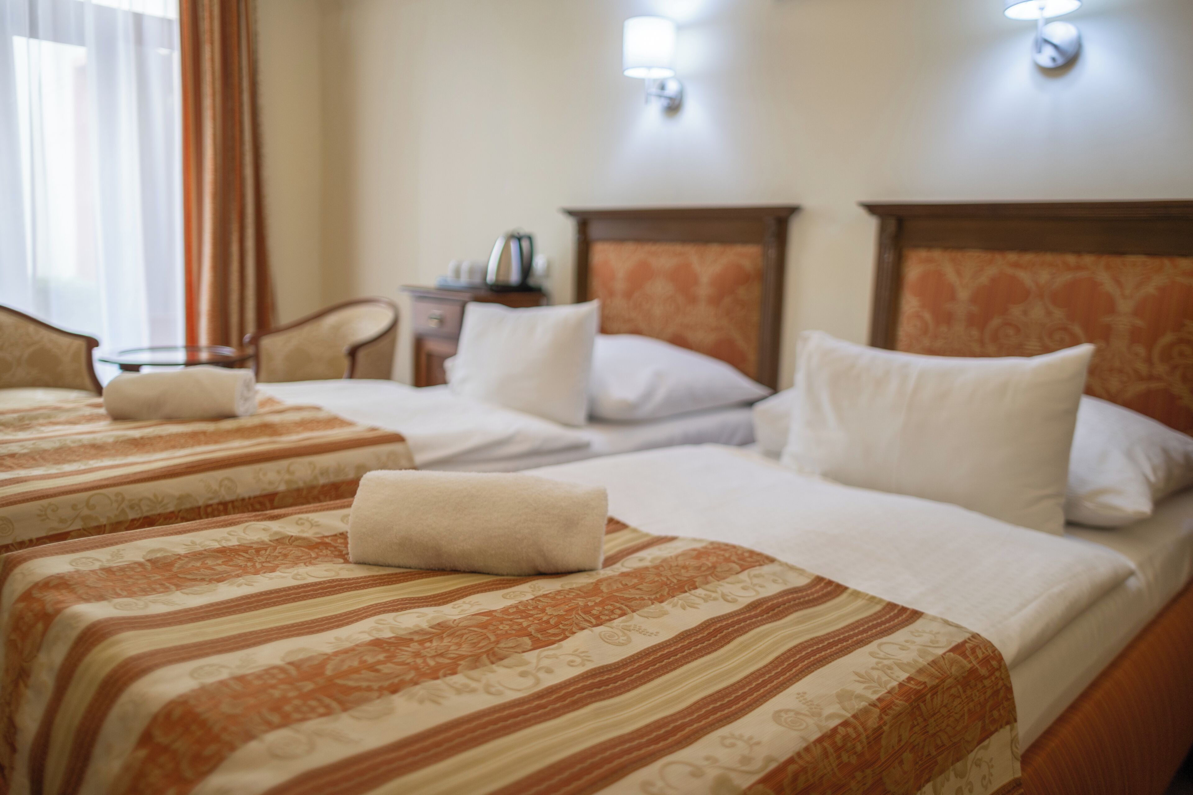 A hotel room with two beds, a nightstand, a lamp, and a window with curtains.