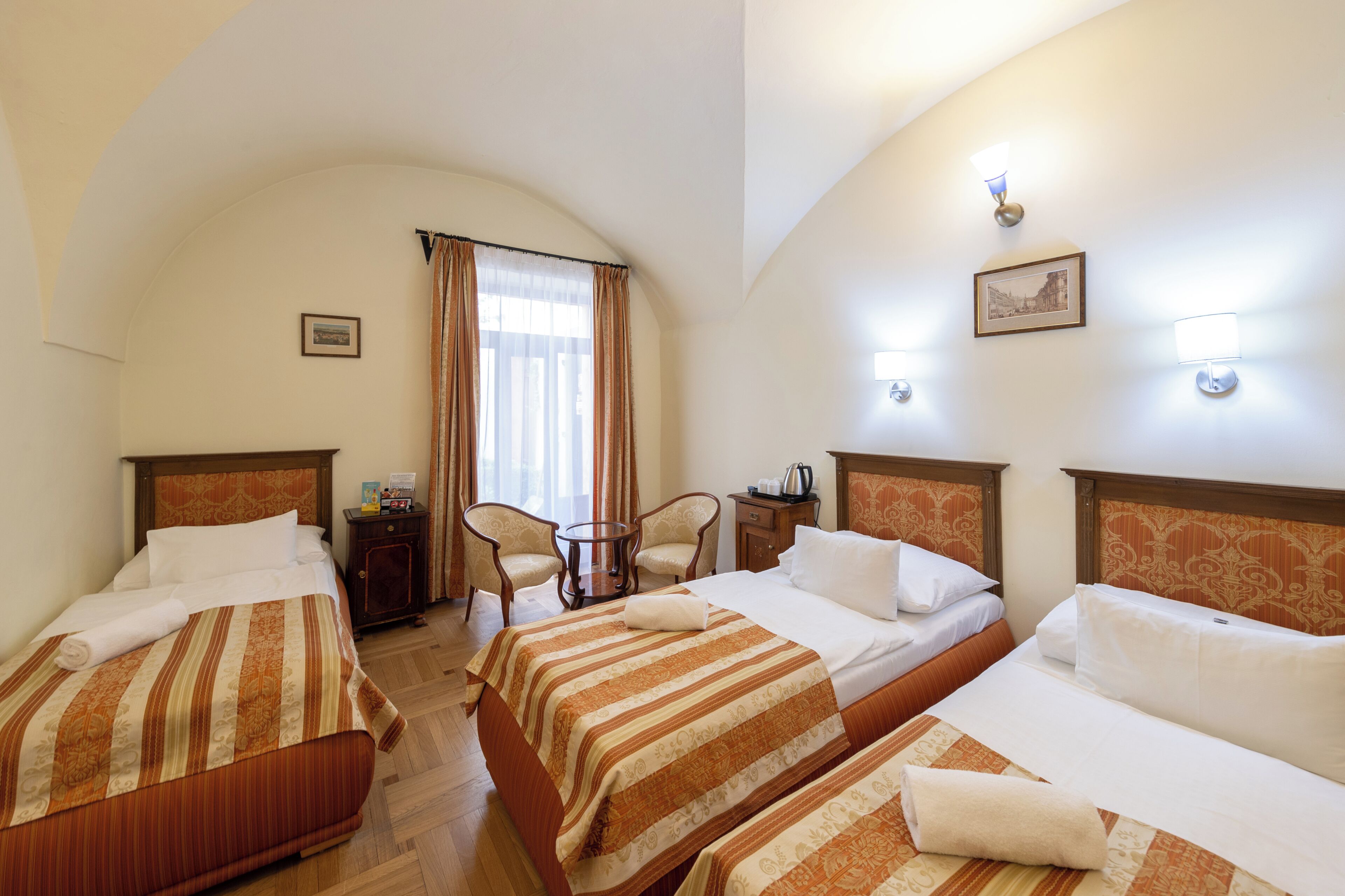 A hotel room with three beds, a small table, two chairs, a window with curtains, and wall-mounted lights.