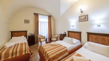 A hotel room with three beds, a small table, two chairs, a window with curtains, and wall-mounted lights.