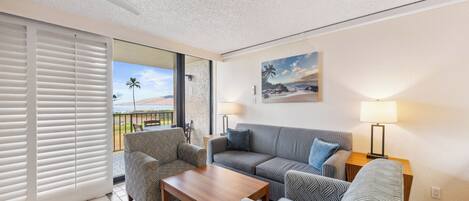 Ein Wohnzimmer mit einer Couch, zwei Sesseln, einem Couchtisch, einem Deckenventilator und Blick auf den Strand durch eine Schiebetür.