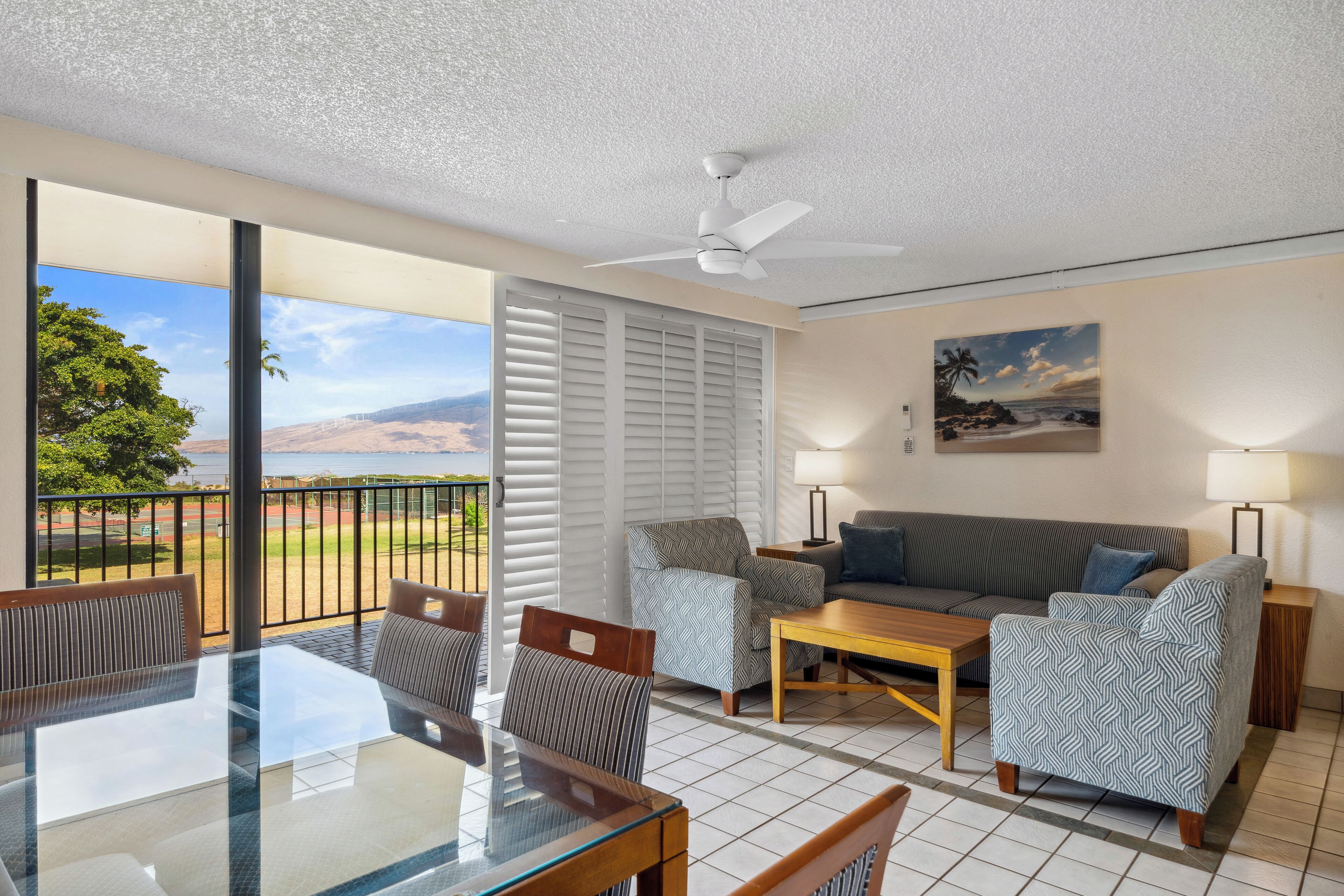 A balcony with a view of the beach and mountains, a glass dining table, a sofa, and patterned armchairs.