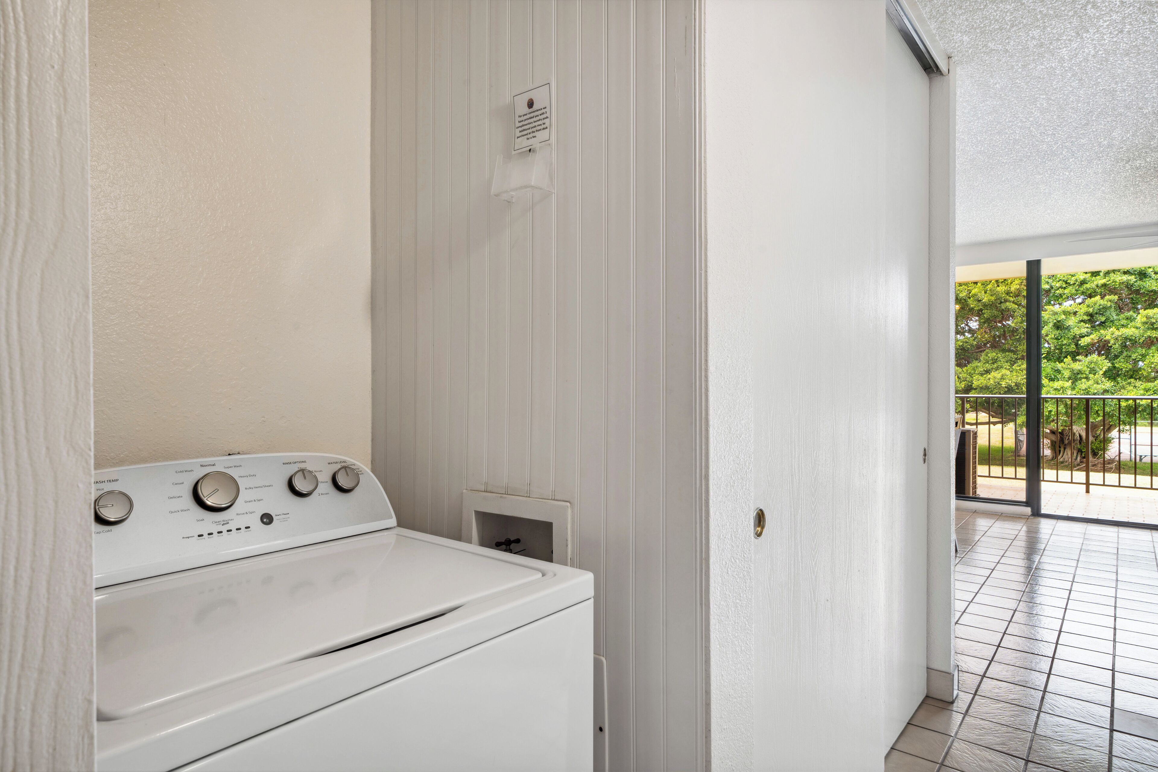 A laundry room with a white washing machine and dryer, a vent, and a door leading to an outdoor balcony with a view of greenery.