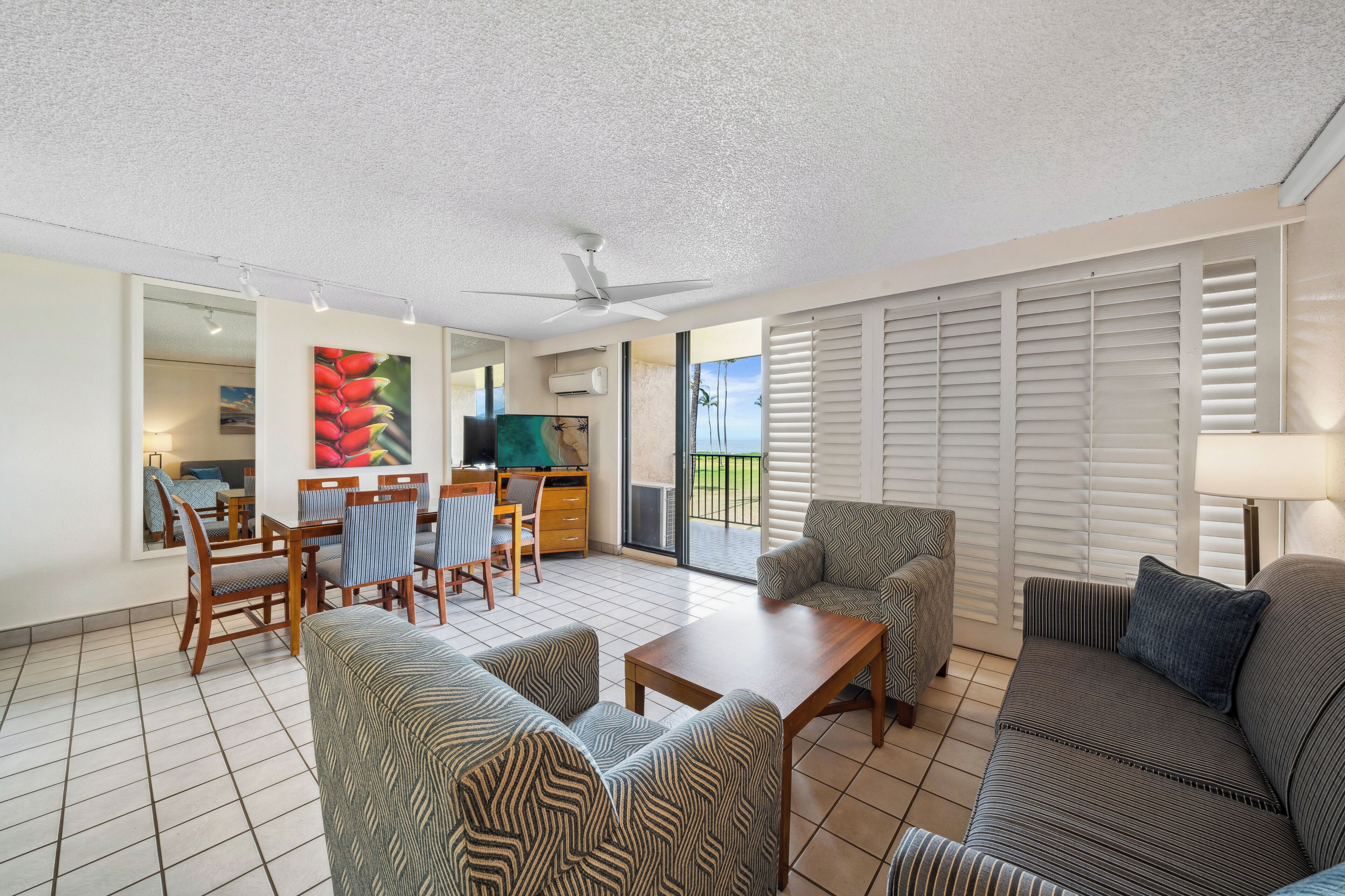 A spacious living area with a sofa, armchairs, a coffee table, and a dining set. There is a ceiling fan, a TV, and a view through the glass door to the outside.