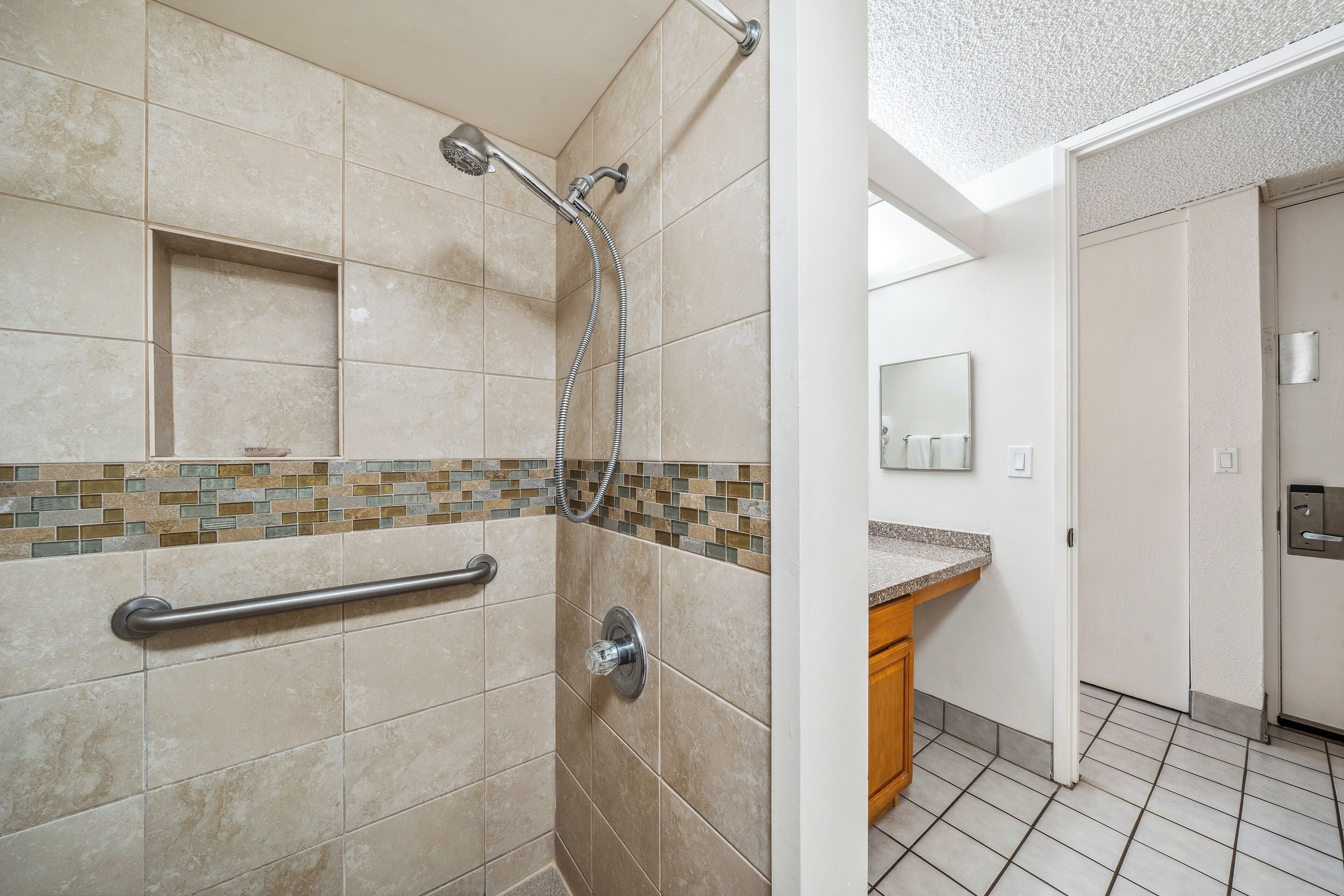 Un baño con baldosas color crema, una ducha con cabezal de ducha manual, un asidero y un salpicadero de azulejos.