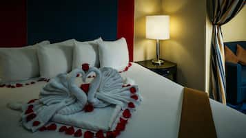 A neatly made bed with white linens and a swan-shaped towel arrangement, red rose petals, and a lamp on a bedside table.