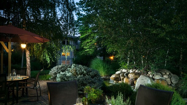 An outdoor dining area with a table set for two, surrounded by chairs, a red umbrella, and a stone water feature.