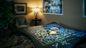 A room with a large puzzle on a table, a lamp, a chair, a plant, and a window with a view of a building.