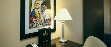 A hotel room with a framed artwork on the wall, a desk with a lamp, a telephone, and a black leather chair.