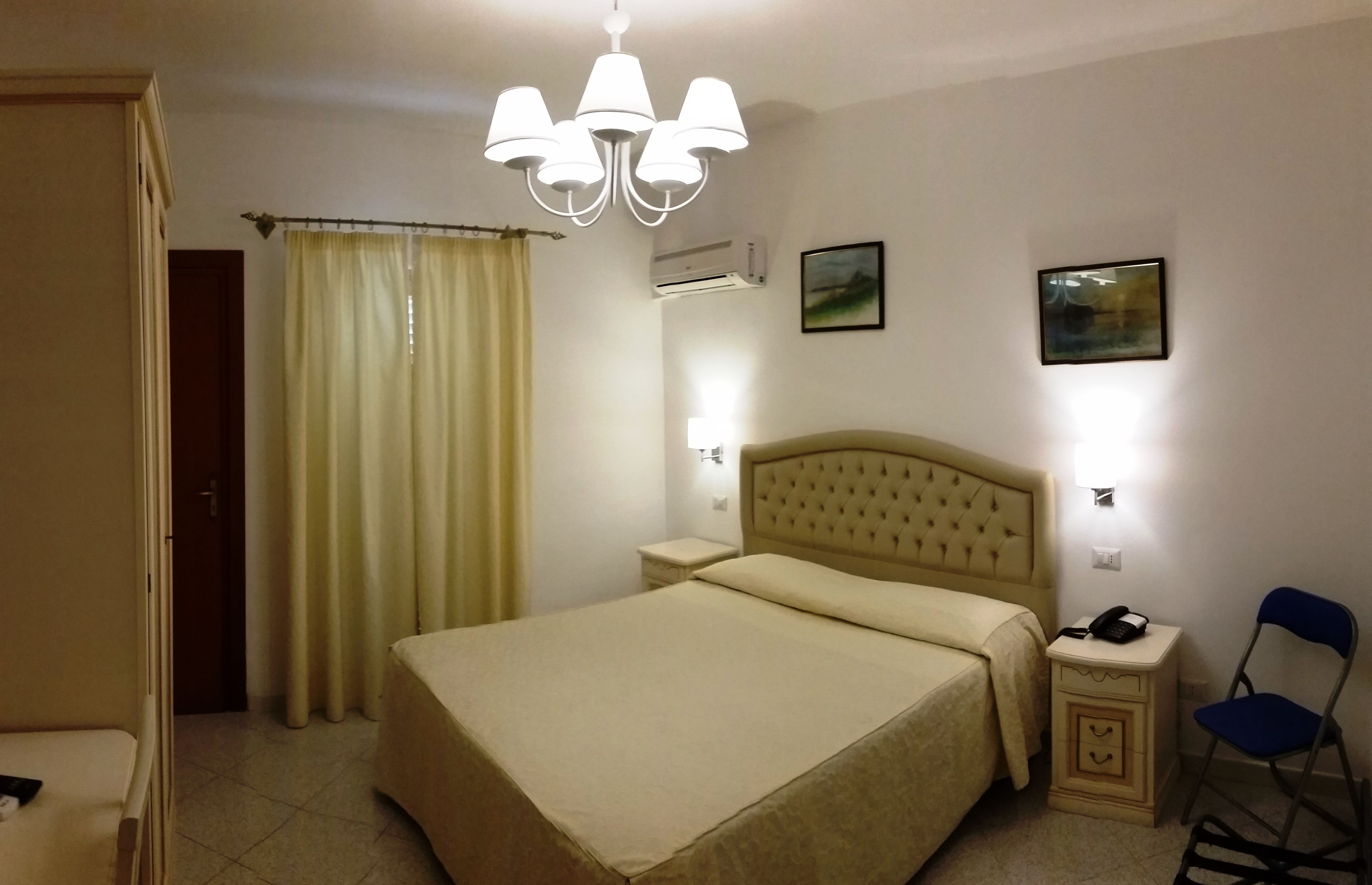 A hotel room with a bed, bedside tables, a chair, a curtain, and a chandelier.
