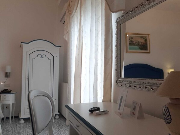 Habitación de hotel con tocador blanco, espejo con marco decorativo, cama con cabecera azul y ventana con cortinas de gasa.