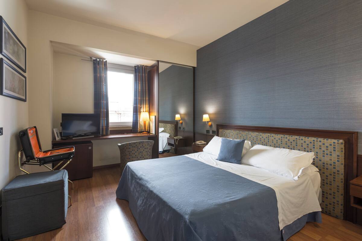 A hotel room with a large bed, a desk with a computer, a chair, a small table, and a mirror.