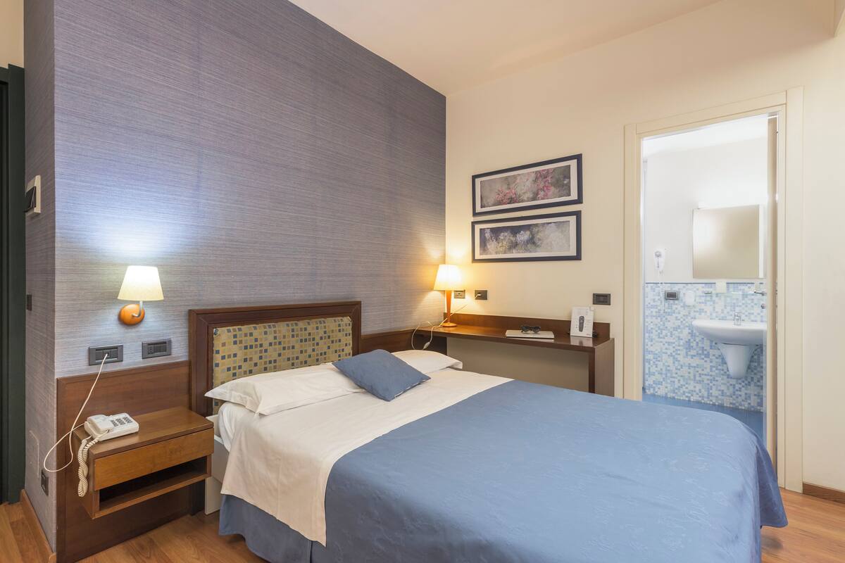 A hotel room with a bed, bedside lamps, a nightstand with a phone, a wall-mounted sink, and framed artwork.
