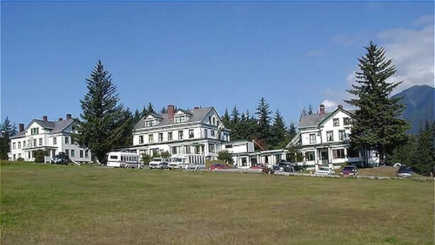 A large, multi-story building with a symmetrical facade, likely a hotel or resort, surrounded by a grassy field and tall trees. - Hotel Halsingland (Haines)